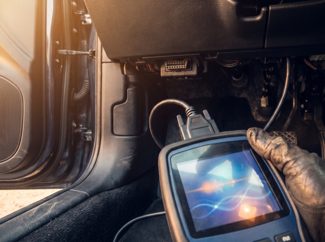 A diagnostic scan tool plugged into the port under a vehicle dashboard, held in a gloved hand